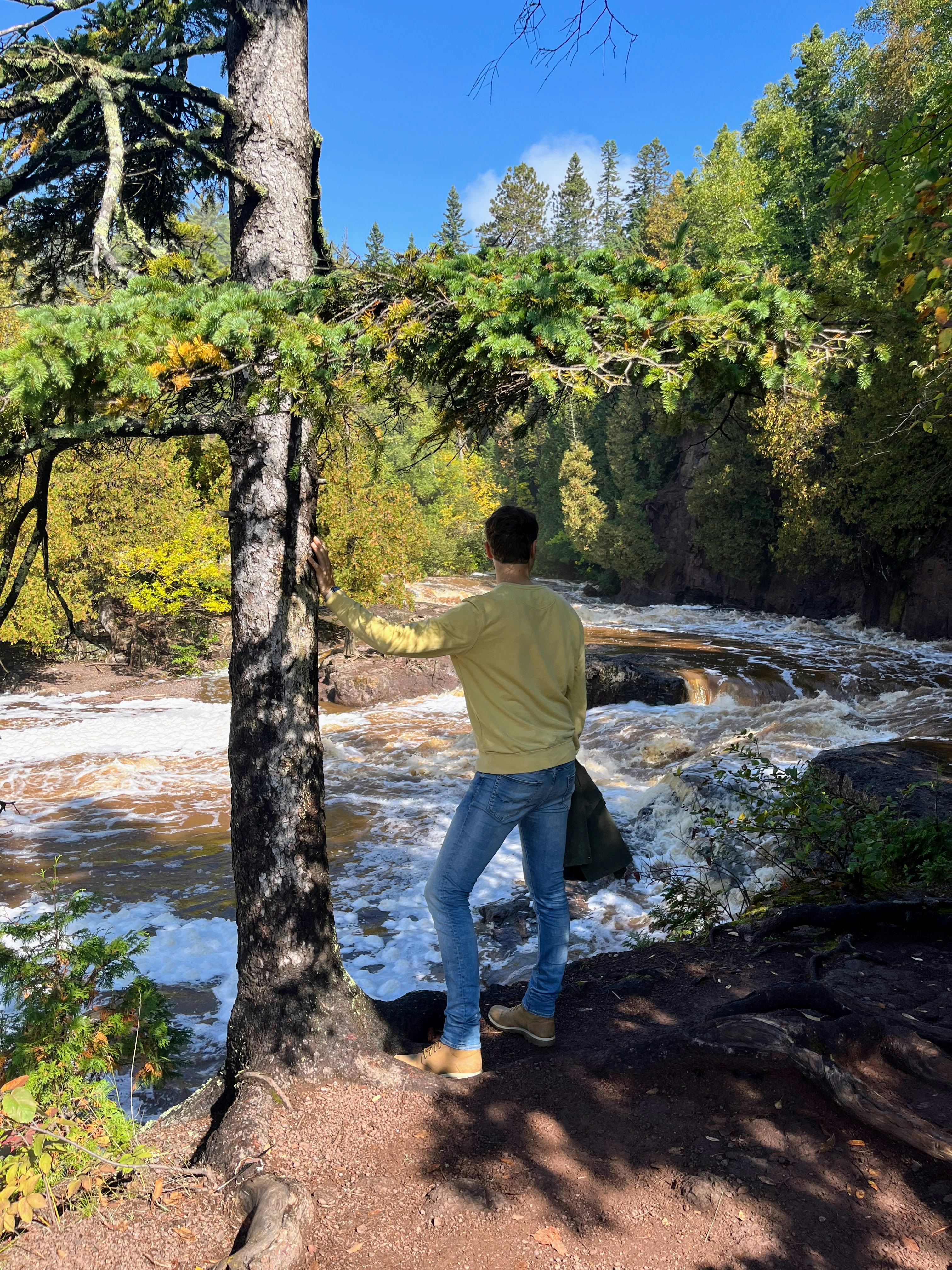 Connor stands next to a pine tree looking down onto the rushing and roaring river below