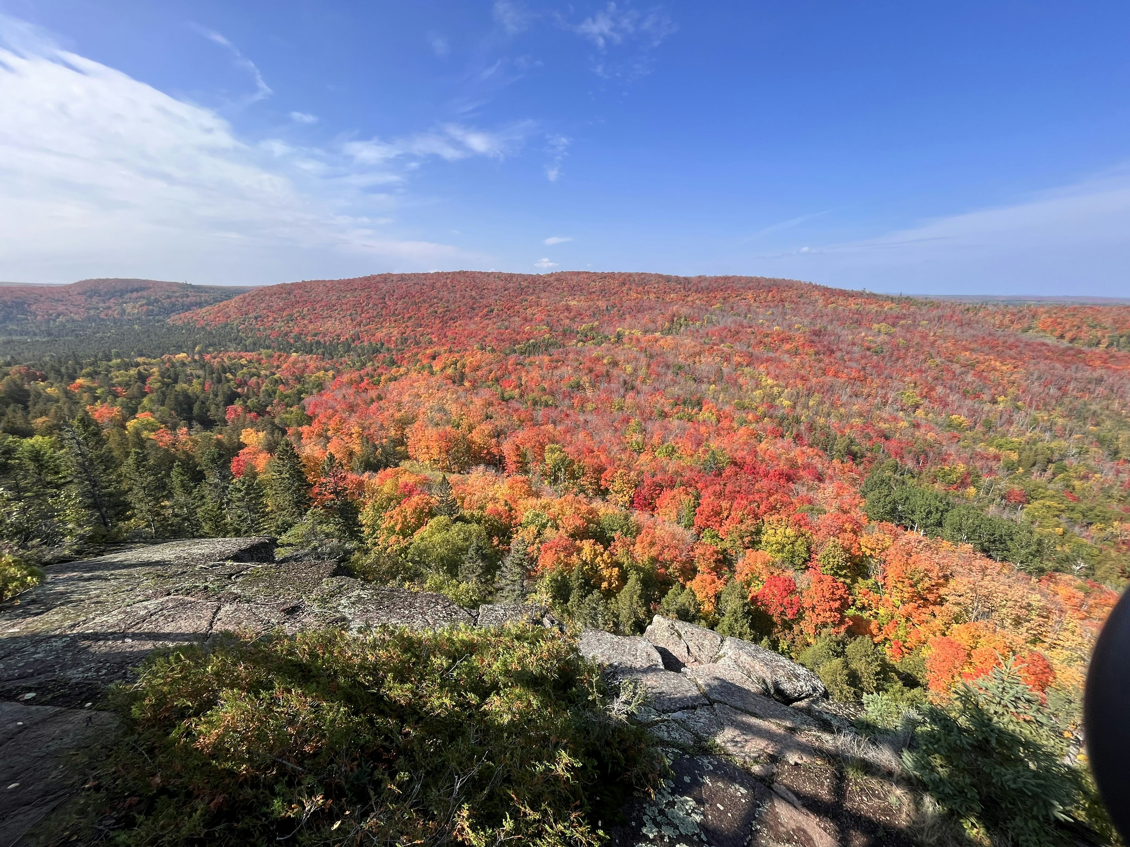 A rocky outlook in the foreground offers a panoramic view of the surrounding fall-colored forest. Bright red, orange and yellow trees are interspersed with green pine trees