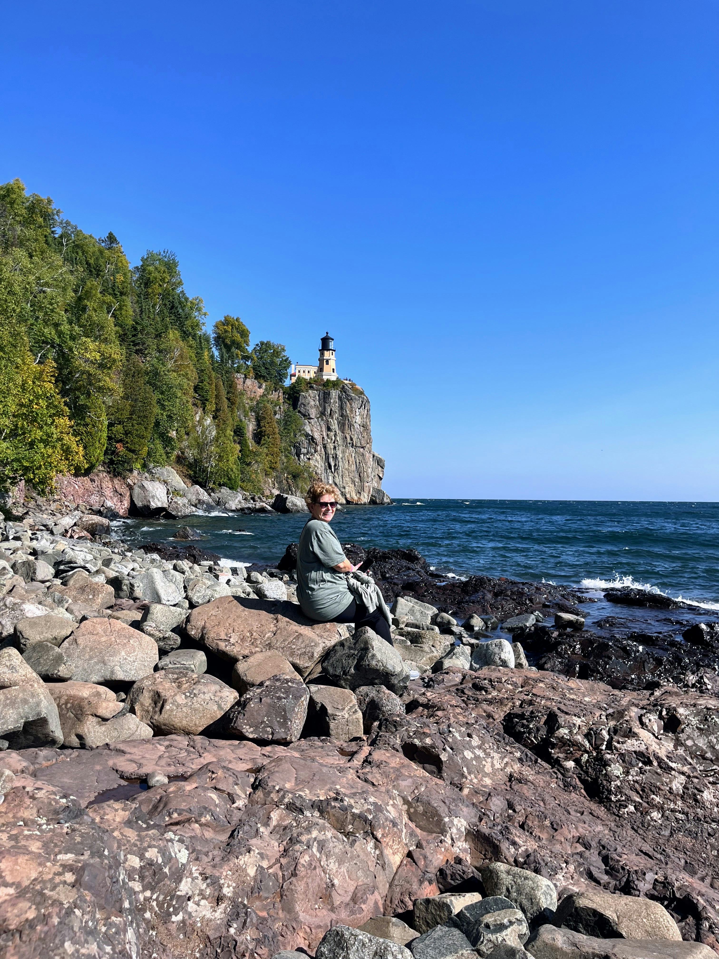 Julie sits in the middle of a rocky beach. In the background, the wooded coast line rises to a towering rock topped by a yellow lighthouse
